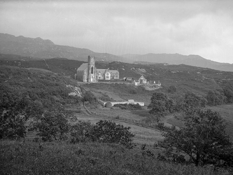 Black and white view of a tall, Gothic-style church set among hills with trees, grass and heather.