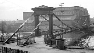Black and white view of St Andrews Suspension Bridge, a pedestian bridge over the River Clyde. In the background stands a large, industrial building.