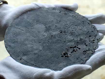 Gloved hands cradling an ancient, fragile-looking disc of lead, its diameter about as long as a hand and its grooved surface pitted with age