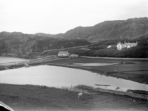 Black and white view showing a pond or partially flooded field, separated from another body of water by a built up road or causeway. Behind the field stands a small boathouse and a larger two-storey house, set among trees.