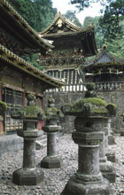 Traditional Japanese stepped temple roofs ornamented with gold, with mossy stone lanterns on the gravel-covered path below