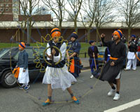 Procession of Sikhs in bright colours and turbans walking along an urban side street. One boy twirls a rope, making circular patterns