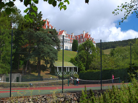 Colour view of a large, four-storey hotel set on a grassy hillside. In the foreground is a group of fenced-off tennis courts, set among trees.