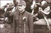 Photograph showing a single S.S. officer at the Belsen camp with corpses piled high on a truck behind him