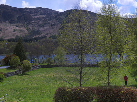 Colour view of an untended garden bounded by stone walls, set next to a narrow loch with tall hills in the background. Aside from a small hedge, some broken-down walls and three tall trees, the garden is given over to grass. To the left, a woman in a red anorak stands on an overgrown path.