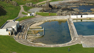 Colour view looking down from coastal cliffs to abandoned open air swimming complex. The pool to the left is partially dried up. The Art Deco pavillion to the left appears partially boarded up.