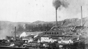 Black and white view of extensive industrial buildings with tall smoking chimney stacks with tall, flat topped hills behind.