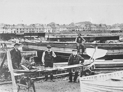 A group of men in Victorian dress stand among boats and carpentry equipment on the shoreline with a row of cottages behind.