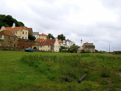 Colour view of a wide, flat expanse of grass with a village rising up the hill behind. The village features a variety of traditional, harled buildings with tile or slate roofs. In the background is a tollbooth with what appears to be one or two mid-20th Century houses in front of it.
