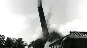 Black and white view of the demolition of a tall, industrial chimney stack.