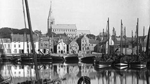 Black and white view of harbour filled with sailed fishing boats with town dominated by church tower in the background