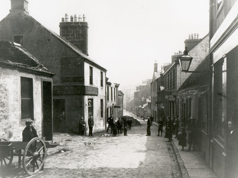 Black and white view down New Street, Paisley. The cobbled street is lined by one and two-storey buildings. A number of people in Edwardian dress, including a man resting against a two-wheeled cart, look towards camera.