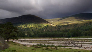 View across winding river on flat valley floor to cloud-darkened hills beyond.