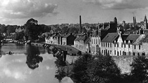 Black and white view of wide flat river with a town to the right of frame. A line of stone, terraced housing lines the street overlooking the river. Several tall industrial chimneys can be seen behind.