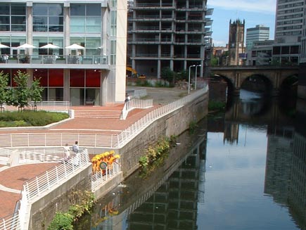 The monks are gathered on a small dock by the riverside, scattering the sand on the water. In the background are Manchester's buildings, including the cathedral