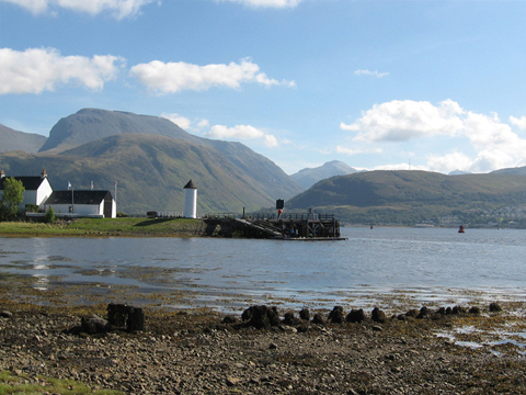 Colour view from a rocky beach across Loch Linnhe to Ben Nevis. In the middle distance is a quayside featuring a cylindrical tower, pitched-roofed buildings and a wooden pier with slipway.
