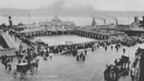 Black and white view of Dunoon Pier lined with crowds of people. Horses and carriages are on the street in the foreground. Two steamers with smoking chimneys are moored in the background.
