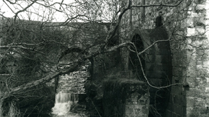 Black and white view of a watermill and wheel next to a narrow burn. An arched stone bridge crosses the burn over a small weir or waterfall.