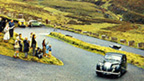 Colour image of a vintage Daimler motorcar climbing a steep, double-hairpin section of road in a mountainous area of grass and heather. A small crowd of people stand waving at the roadside.