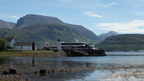 Colour view from a rocky beach across Loch Linnhe to Ben Nevis. In the middle distance is a quayside featuring a cylindrical tower, pitched-roofed buildings and a wooden pier with slipway.