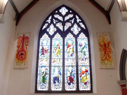 A tall stained glass window designed by William Morris, depicting eight saints.  On either side of the window are hangings showing a person with the flame of the Holy Spirit, captioned 'Prayer'