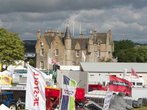 Colour view of large Scottish Baronial house. In the foreground are the temporary structures and banners of an agricultural show.