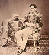 Emperor Meiji, a Japanese man in formal military dress, sits back in a fancy antique chair.  A fringed hat like that of a military commander can be seen on the table next to him and the upholstery, carpet and tablecloth are richly patterned