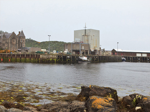 Colour view across water to a harbour, where a small fishing boat is tied up. The harbour features a number of industrial-scale modern buildings. The large house to the left of frame has been restored to use.