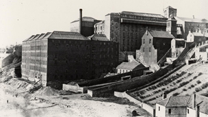 Black and white view of extensive factory building, set on a slope leading down to a beach at the bottom left of frame.