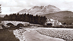Black and white view of a braided river crossed by a twin-arched road bridge. Behind stands the Kinlochewe Hotel with the mountain Sgurr Ban in the background.