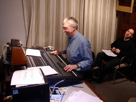 Sound engineer and producer sitting by the sound desk, with formidable rows of switches and sliders. They are in a small room, watching the service on a monitor in the foreground