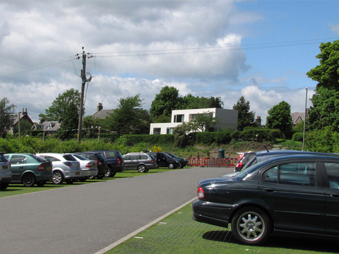Car park in rural setting with white modernist building behind.