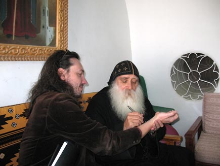 Father Ruwais, a Coptic monk wearing black robes and skull cap and a long white beard, sits drawing on Nicholas Buxton's arm. The monastery room contains wooden furniture and a round patterned glass window with a painting of a saint visible on one of the white-painted walls