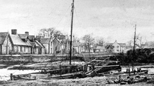 Black and white view of sailing boat in small harbour with row of cottages behind.