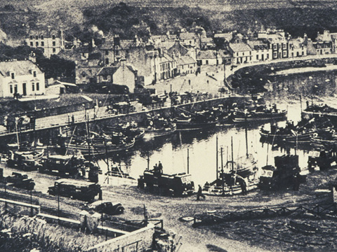 Black and white view of harbour with fleet of fishing boats and a number of loaded trucks on the quayside.