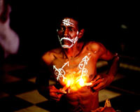 Shirtless dark-skinned man with symbols drawn on his face and body in bright white, holding lit candles against his chest