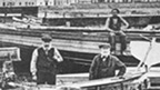 A group of men in Victorian dress stand among boats and carpentry equipment on the shoreline with a row of cottages behind.