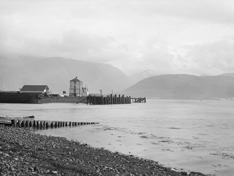 Black and white view from a rocky beach across Loch Linnhe to Ben Nevis. In the middle distance is a quayside featuring a tall, masonry constructed building and a wooden pier.