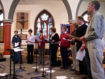 The Daily Service Singers, nine men and women wearing ordinary clothes, stand in a semicircle around small microphone stands inside the church.  Christian-themed wall hangings and stained glass windows can be seen in the background