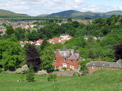 View over town featuring a large Victorian house in the foreground. Behind this, among trees, is a modern group of houses with the main part of the town behind.