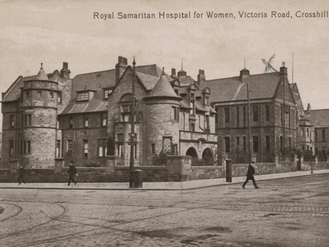 Black and white view of two-storey hospital building featuring a range of pitched-roofed main blocks with turrets. The hospital stands at the crossroads of two wide streets along which run a number of tram tracks.