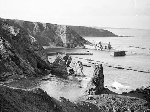 Black and white view looking down from cliffs to small harbour with buildings at Cove.