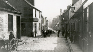 Black and white view down New Street, Paisley. The cobbled street is lined by one and two-storey buildings. A number of people in Edwardian dress, including a man resting against a two-wheeled cart, look towards camera.