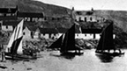 Black and white view of small sailing boats landing on a beach. A cluster of cottages rises up the hill behind.