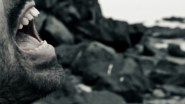 An open human mouth with teeth bared angrily with a rocky background.