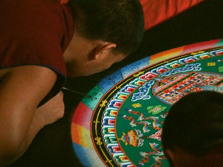 A monk is bending over the outer border, made from stripes of coloured sand, adding intricate detail in black sand