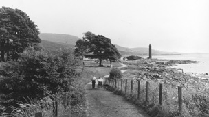 Black and white view of a couple with a pram and dog walking along a winding, coastal footpath. In the background are trees and the tall, cylindrical Battle of Largs Monument.