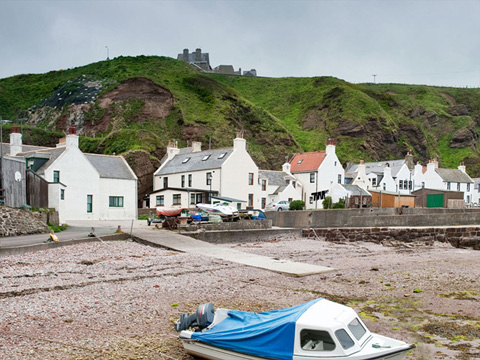 Colour view showing one small motorboat pulled up onto the shore, with a row of cottages beneath tall cliffs behind.