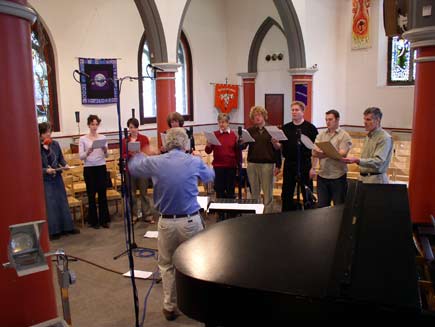 Nigel Swinford conducting nine choir members, grouped in a small floorspace in the middle of the church.  Around them are chairs, microphone stands, an overhead projector and a piano