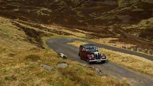 Colour view of a vintage Daimler motorcar climbing a straight section of road with a hairpin bend behind.